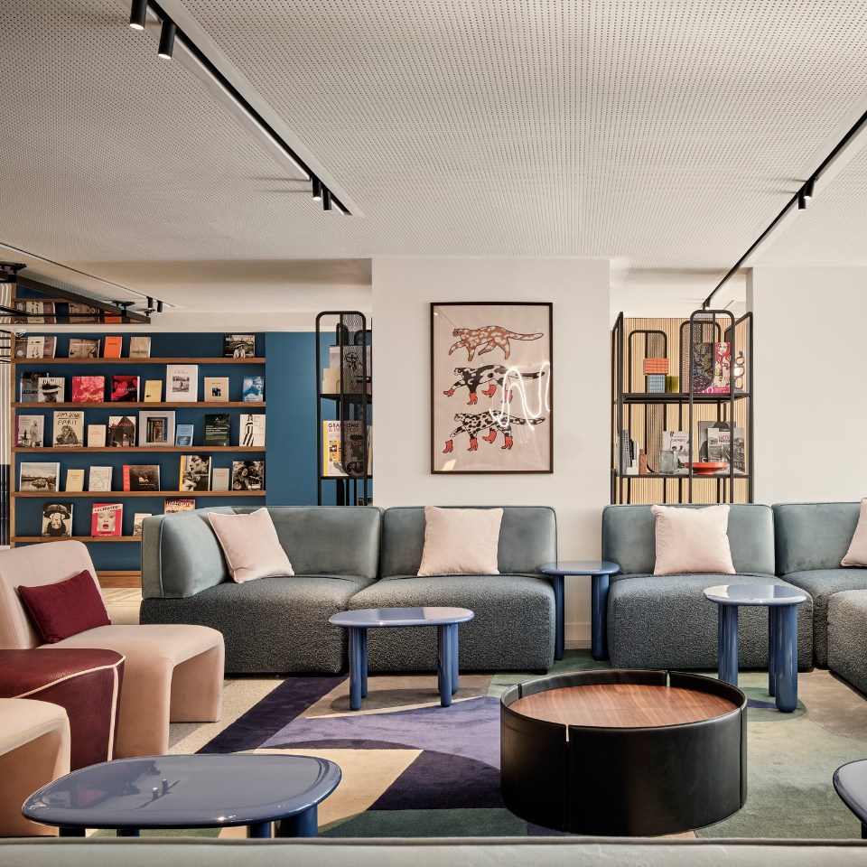 Modern lounge with plush sofas, pastel cushions, and sleek blue tables. A colorful bookshelf and artwork create a cozy, stylish atmosphere.