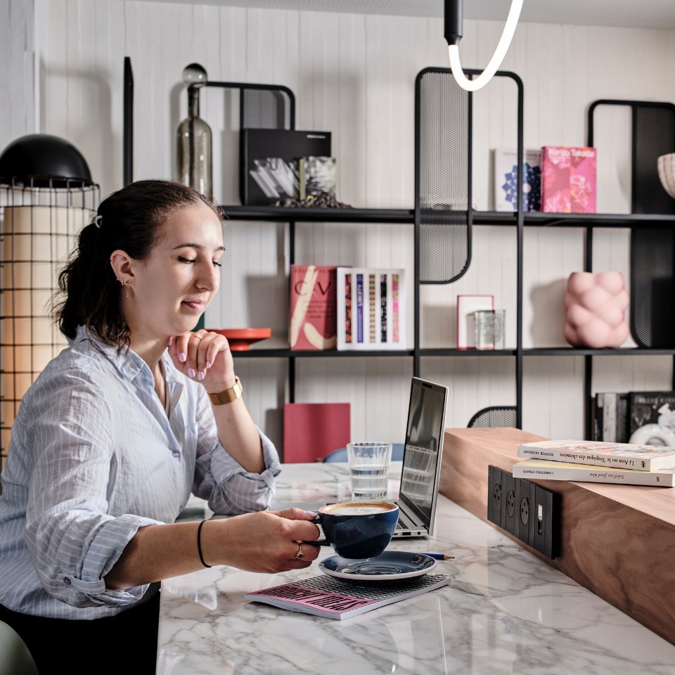 A woman in a striped shirt sits at a marble counter in a modern, cozy space. She holds a coffee cup, with a laptop and books nearby. Shelves with art and decor are in the background.