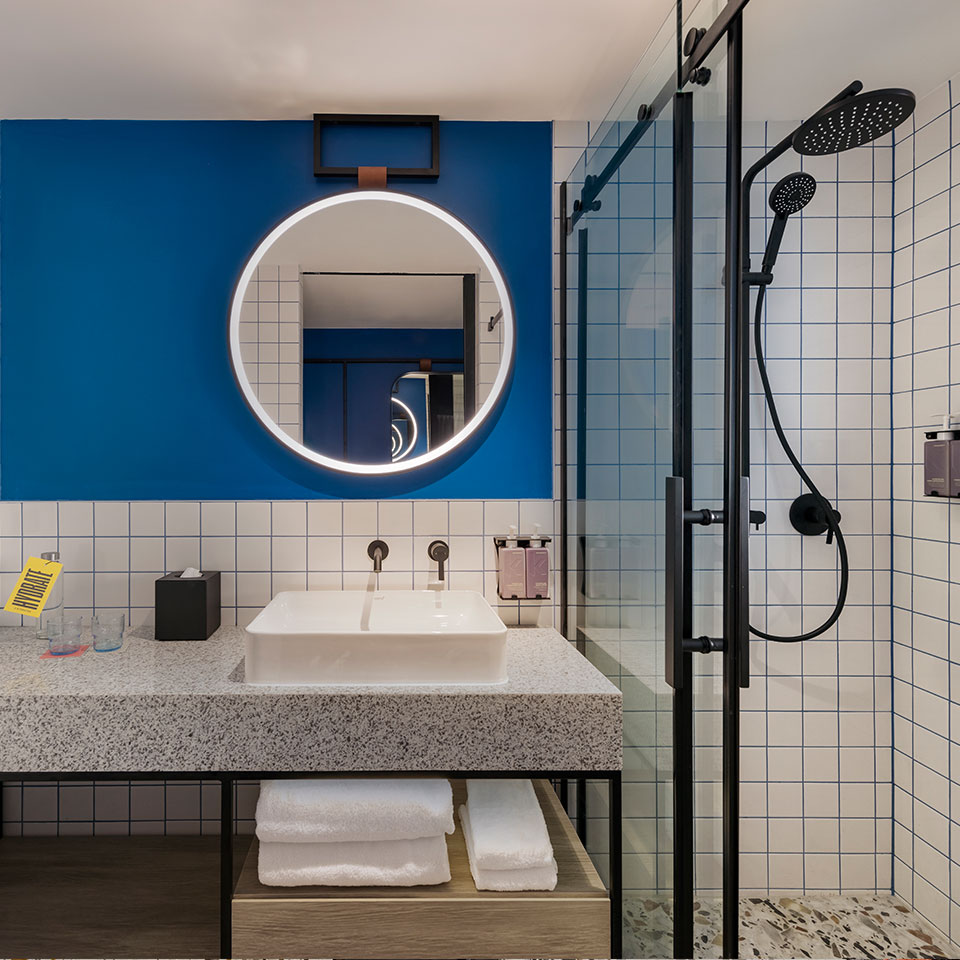 Stylish bathroom with modern round mirror on a blue wall, illuminated by an LED light. Features include a sleek countertop sink, glass shower, and white towels.