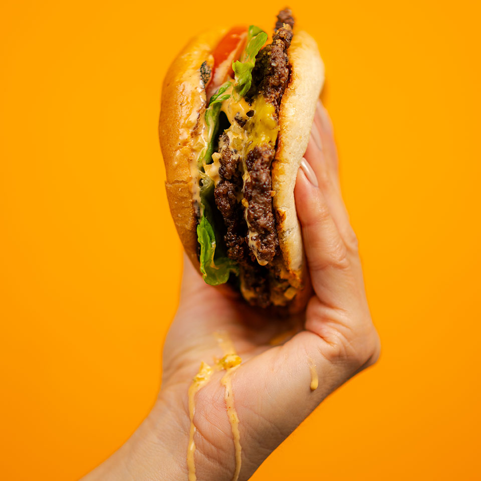 A hand holds a juicy burger with lettuce, tomato, and melted cheese against a vibrant orange background. Cheese drips down the fingers, adding a messy, appetizing touch.