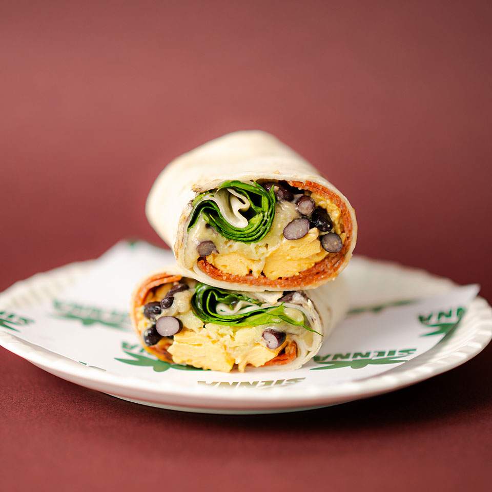 A neatly wrapped burrito cut in half on a white plate against a brown background. Inside are visible eggs, spinach, and black beans, suggesting a hearty, fresh meal.