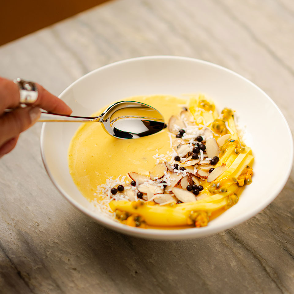 A hand holds a spoon poised over a creamy mango smoothie bowl topped with sliced almonds, coconut, and berries, on a gray marble surface.
