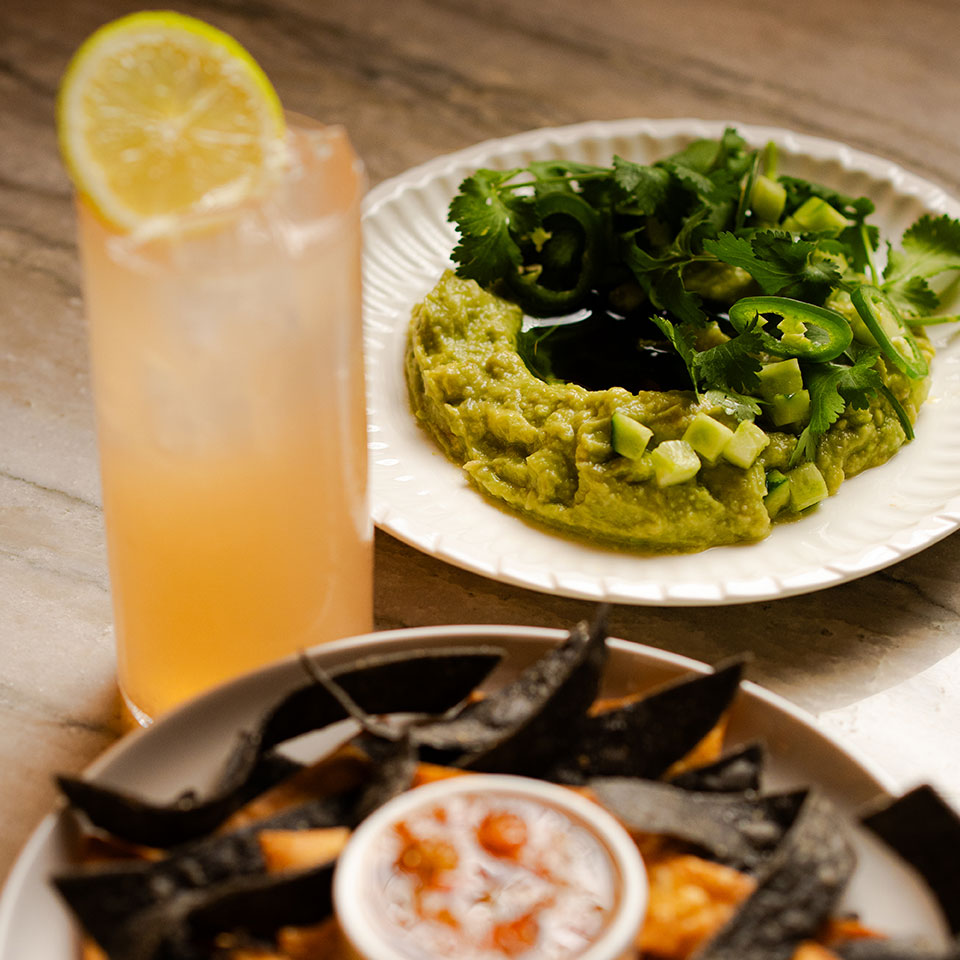 A refreshing lemonade with a lemon slice sits next to a plate of guacamole topped with jalapeños, cilantro, and cucumber, exuding a fresh and zesty vibe.