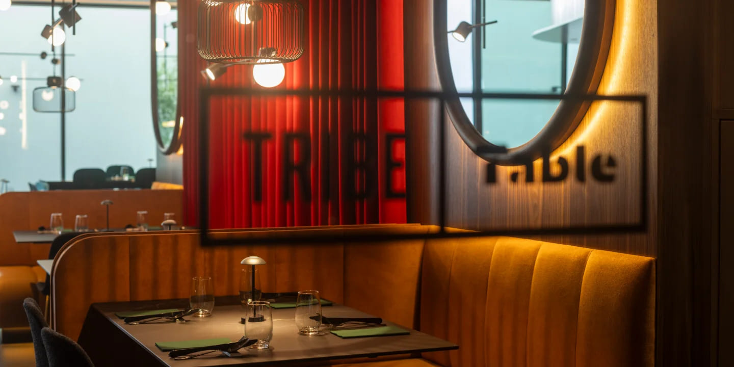 Scène de restaurant chaleureuse avec une décoration moderne, des assises éclairées et une grande enseigne « TRIBE » projetant son ombre sur un mur en bois.
