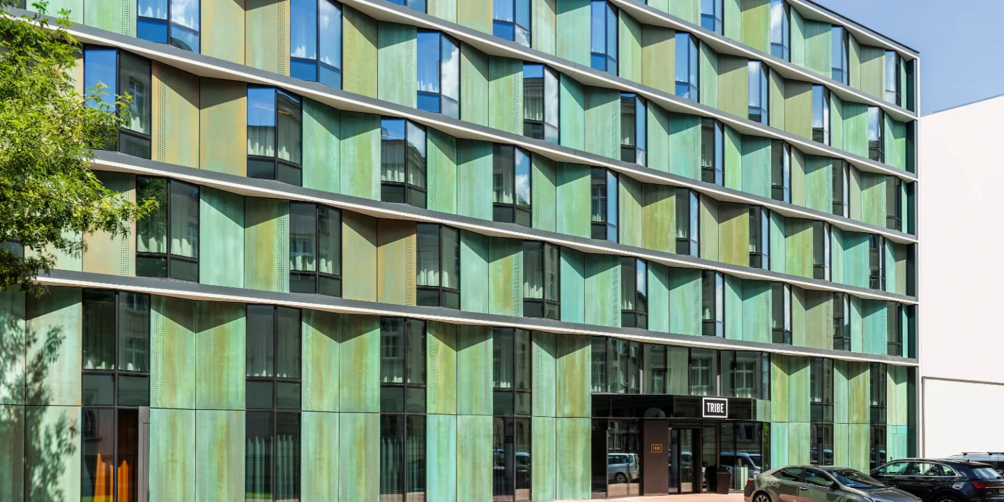 Façade de bâtiment moderne avec des panneaux métalliques verts et de grandes fenêtres, ornée de l’enseigne « TRIBE » à l’entrée. Des voitures sont garées à proximité.