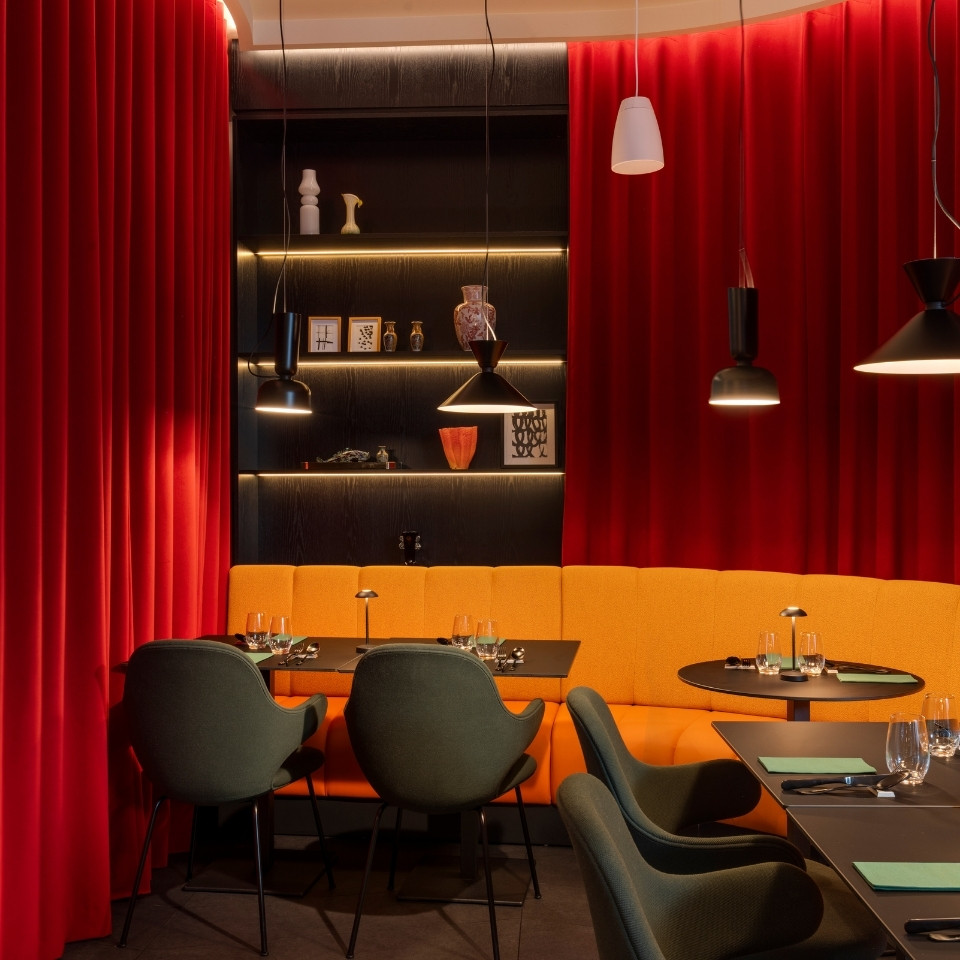 Intérieur de restaurant chaleureux avec des rideaux rouge vif, un coin banquette orange et d’élégantes tables noires à l’éclairage moderne.