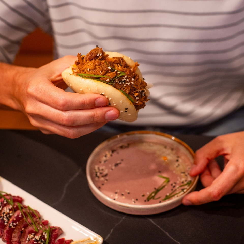 Un homme tient un sandwich d’une main, avec un petit bol de sauce posé à côté sur une table.