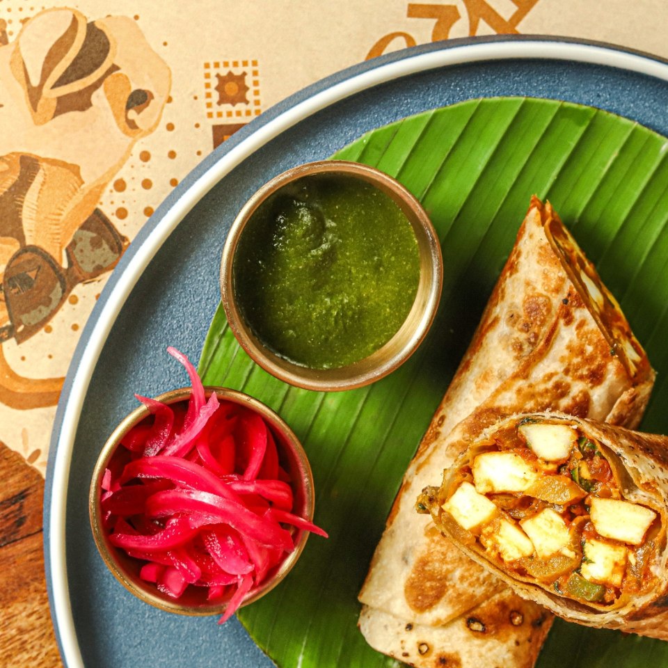 Une assiette colorée avec un wrap au paneer sur une feuille de bananier, accompagné de chutney vert et d’oignons marinés. L’ensemble dégage une ambiance fraîche et colorée.