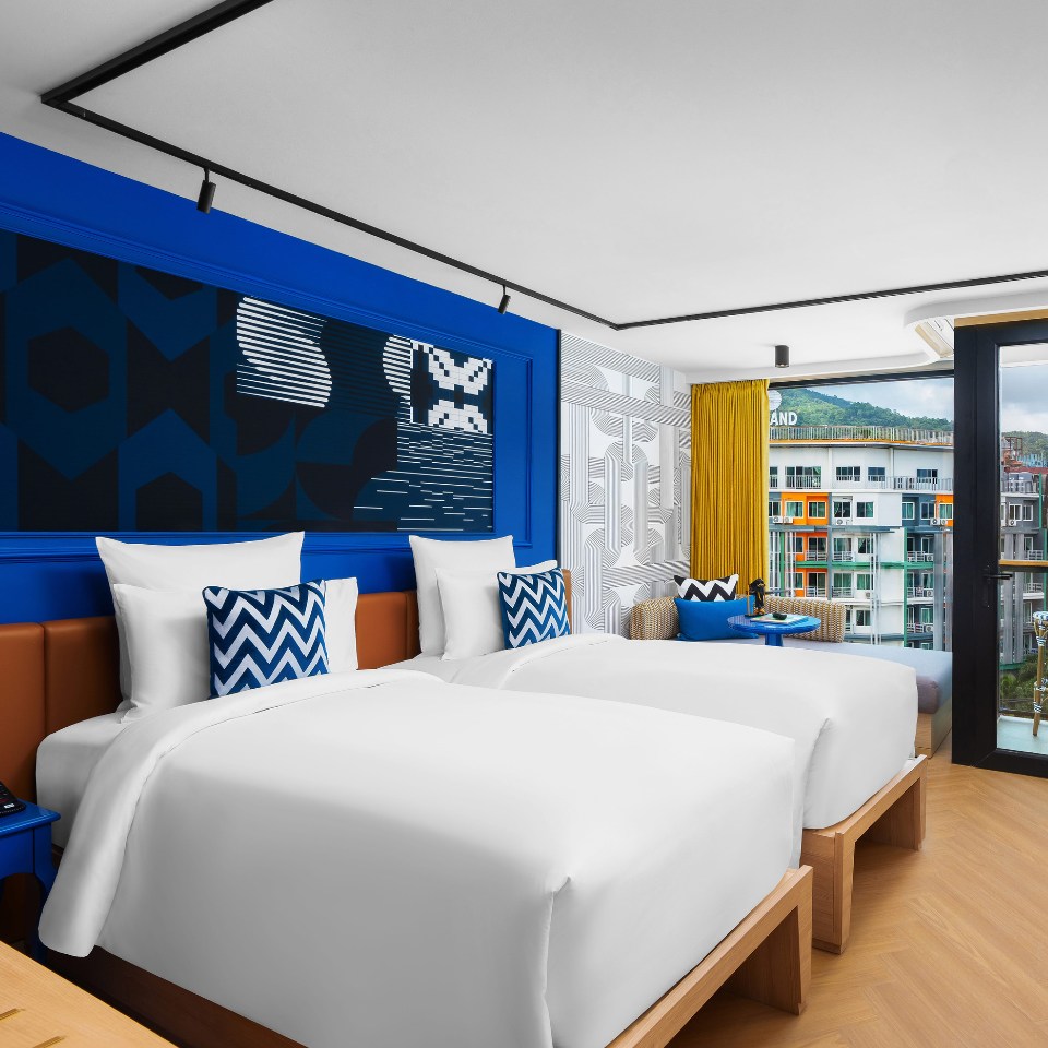 Modern hotel room with two twin beds, white bedding, blue and geometric accents. A balcony reveals a view of colorful buildings and hills. Cozy and vibrant.