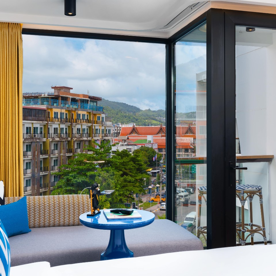 Room with large windows overlooks urban scene with colorful buildings, green hills, and cloudy sky. Cozy sofa, blue table, and vibrant curtains inside.