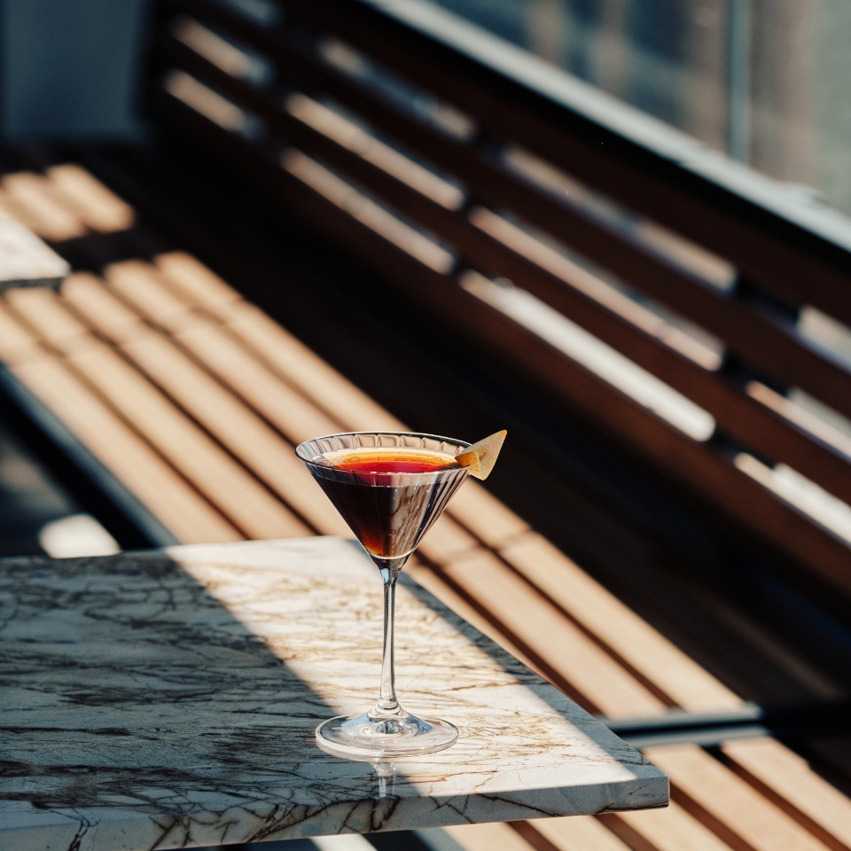 A martini glass with a dark beverage and an orange twist sits on a marble table. Sunlight creates bold shadows and a warm, inviting atmosphere.