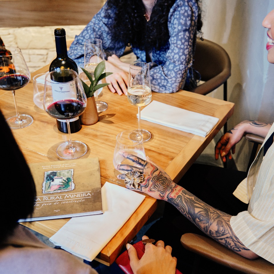 A group of people seated at a wooden table with wine glasses and a menu. One person has a tattooed arm and holds a glass, the setting is cozy and social.