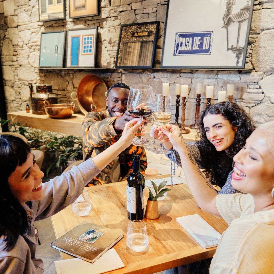 Four friends joyfully clink wine glasses around a wooden table in a cozy restaurant with stone walls and eclectic framed art. Smiles and warmth fill the scene.
