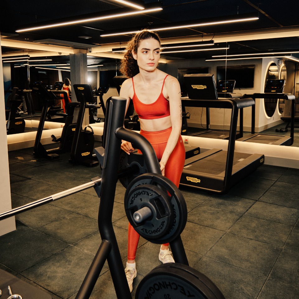 A person in a red workout outfit stands beside a barbell rack in a modern gym. Treadmills are visible in the background, reflecting a focused and determined atmosphere.