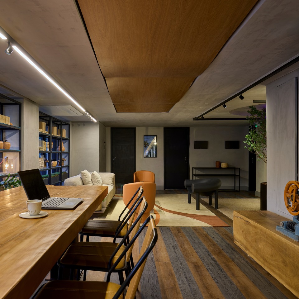 A modern, stylish office space with a long wooden table, four chairs, and a laptop. A cozy sofa, orange chair, and bookshelf create a warm, inviting atmosphere.