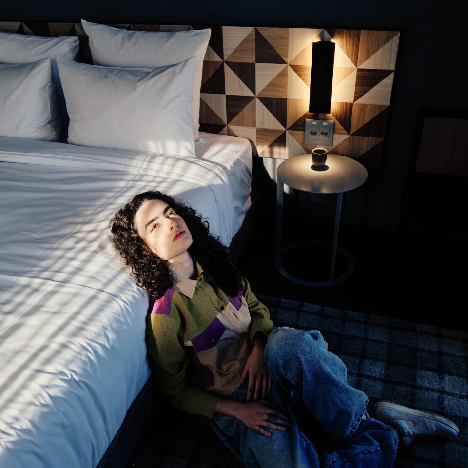 A person seated on a patterned rug beside a bed, dressed in a colorful shirt and jeans, with a bedside lamp casting soft light.