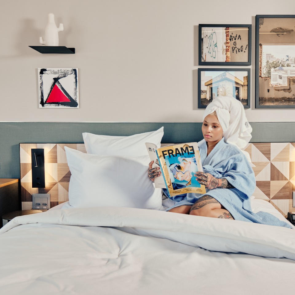 A person in a bathrobe sits on a bed, reading a magazine titled "FRAME," surrounded by modern decor and artwork.