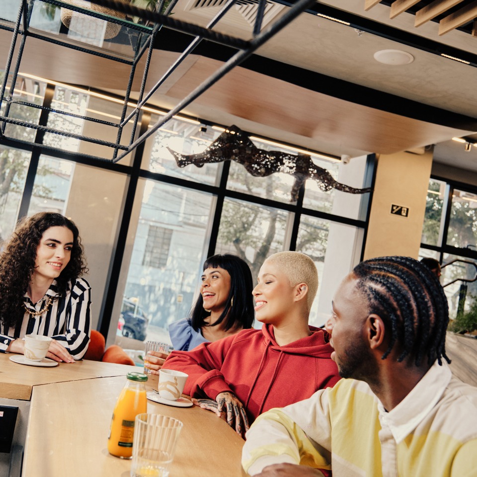 A group of friends enjoying drinks and conversation around a stylish café table, with large windows letting in natural light.