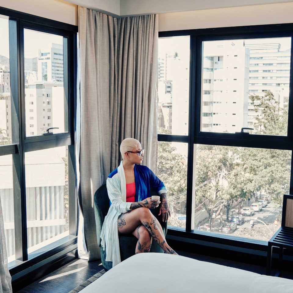 A person in a blue robe sits by a large window, holding a cup, with a cityscape visible in the background.