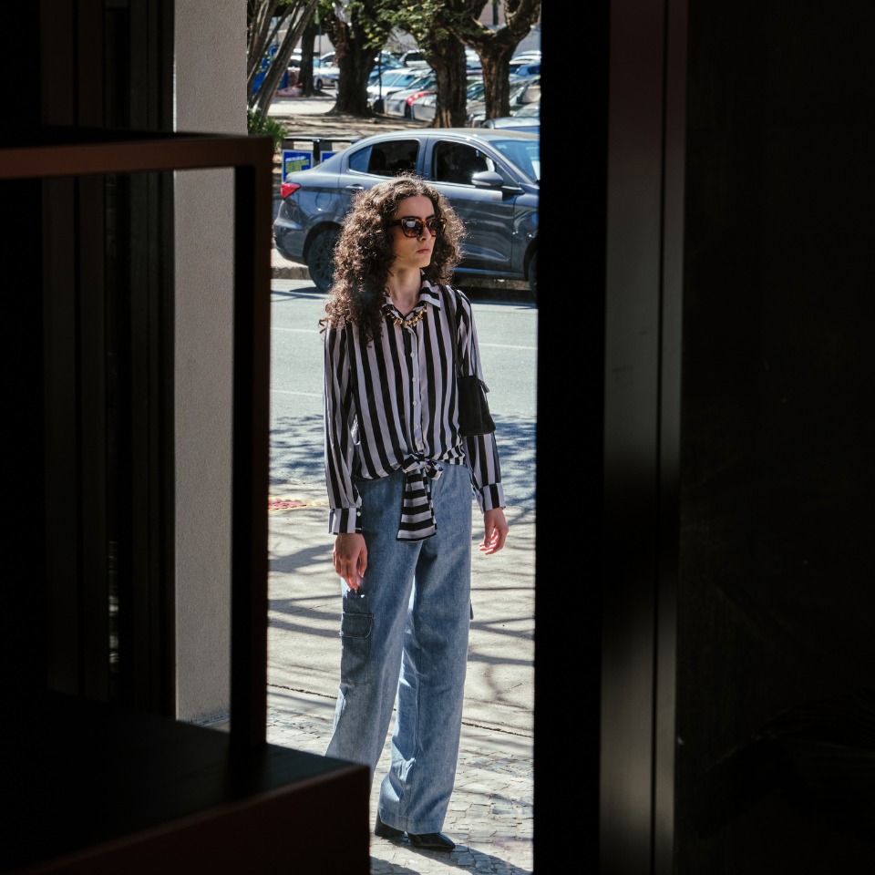 A person in a striped shirt and wide-leg jeans stands at an entrance, framed by a doorway, with trees and cars in the background.
