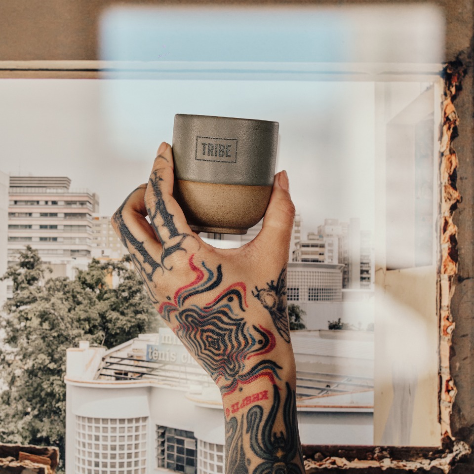 A tattooed hand holds a ceramic cup labeled "TRIBE," with a cityscape visible through a window in the background.