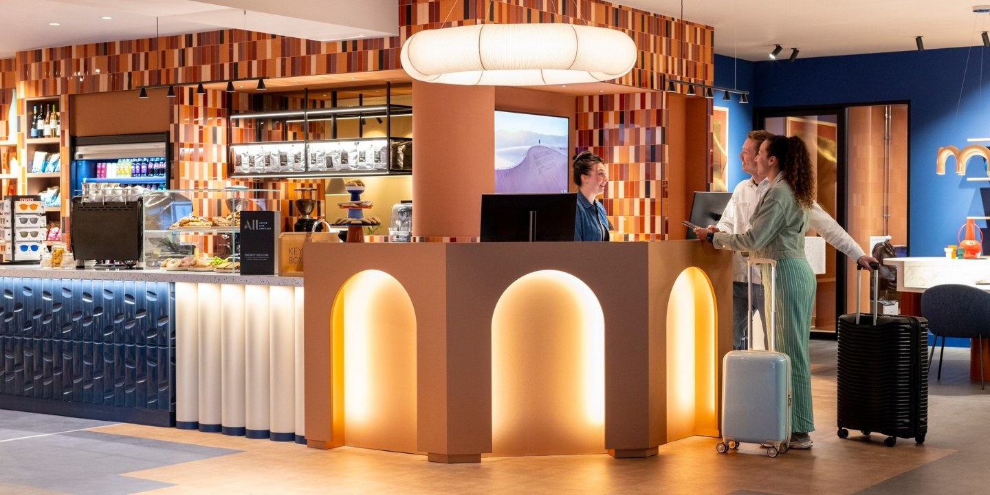 Modern hotel reception area with colorful decor, featuring a service counter, food display, and a traveler with luggage.