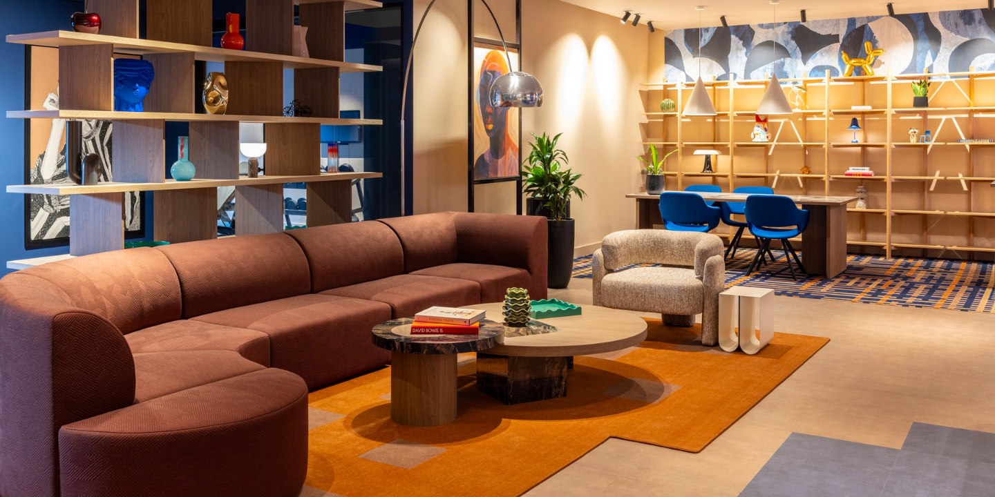 Modern living room with a brown curved sofa, orange rug, abstract art, and plants. Shelving and a cozy seating area create a vibrant, stylish ambiance.