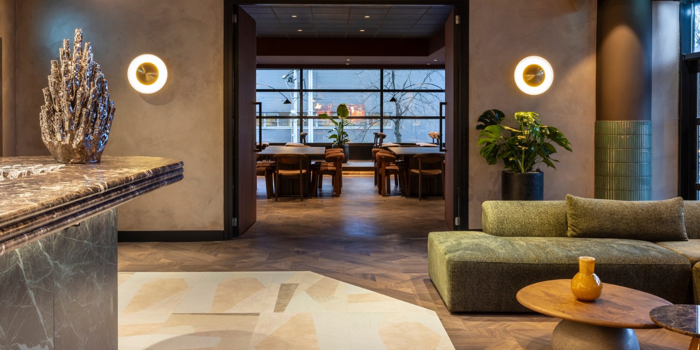 Spacious modern lounge with a marble counter, plush green sofa, and round tables. Warm lighting, large plant decor, and a distant dining area evoke coziness.