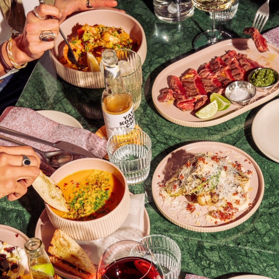 A vibrant table spread features dishes like soup with bread, grilled steak, and a colorful salad, surrounded by drinks and tableware.