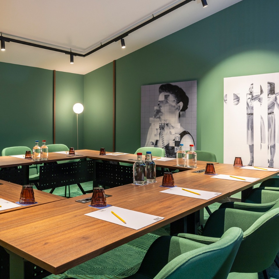 Modern conference room with U-shaped wooden table, green chairs, and minimal decor. Art on green walls; bottled water and notepads neatly arranged.