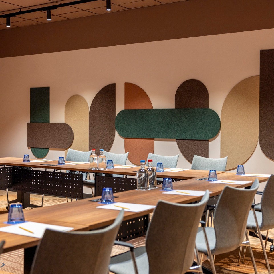 Modern conference room with wooden tables and gray chairs, featuring colorful geometric wall art. Blue glasses and notepads are neatly arranged.