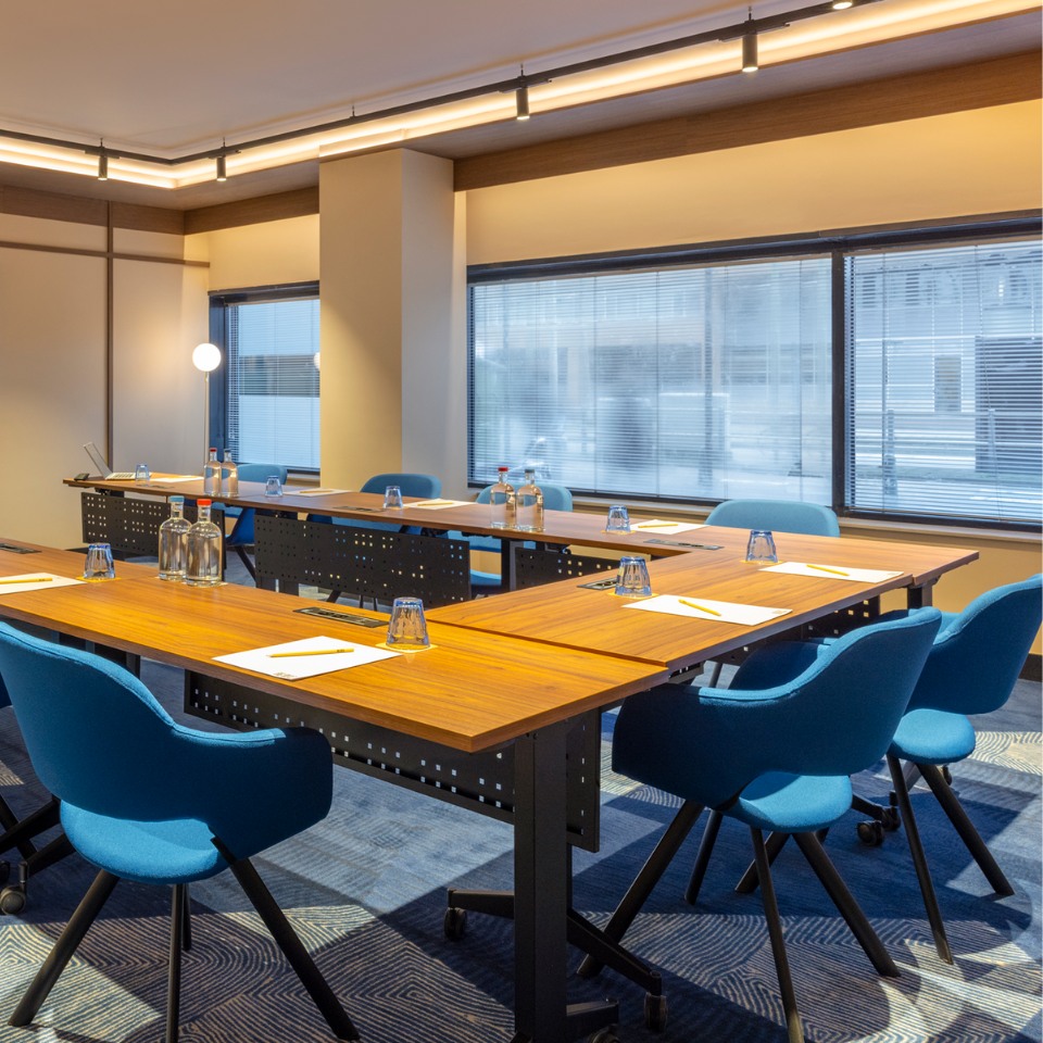 Modern conference room with U-shaped setup. Wooden tables, blue chairs, notepads, and water bottles on tables. Bright, with large windows and soft lighting.
