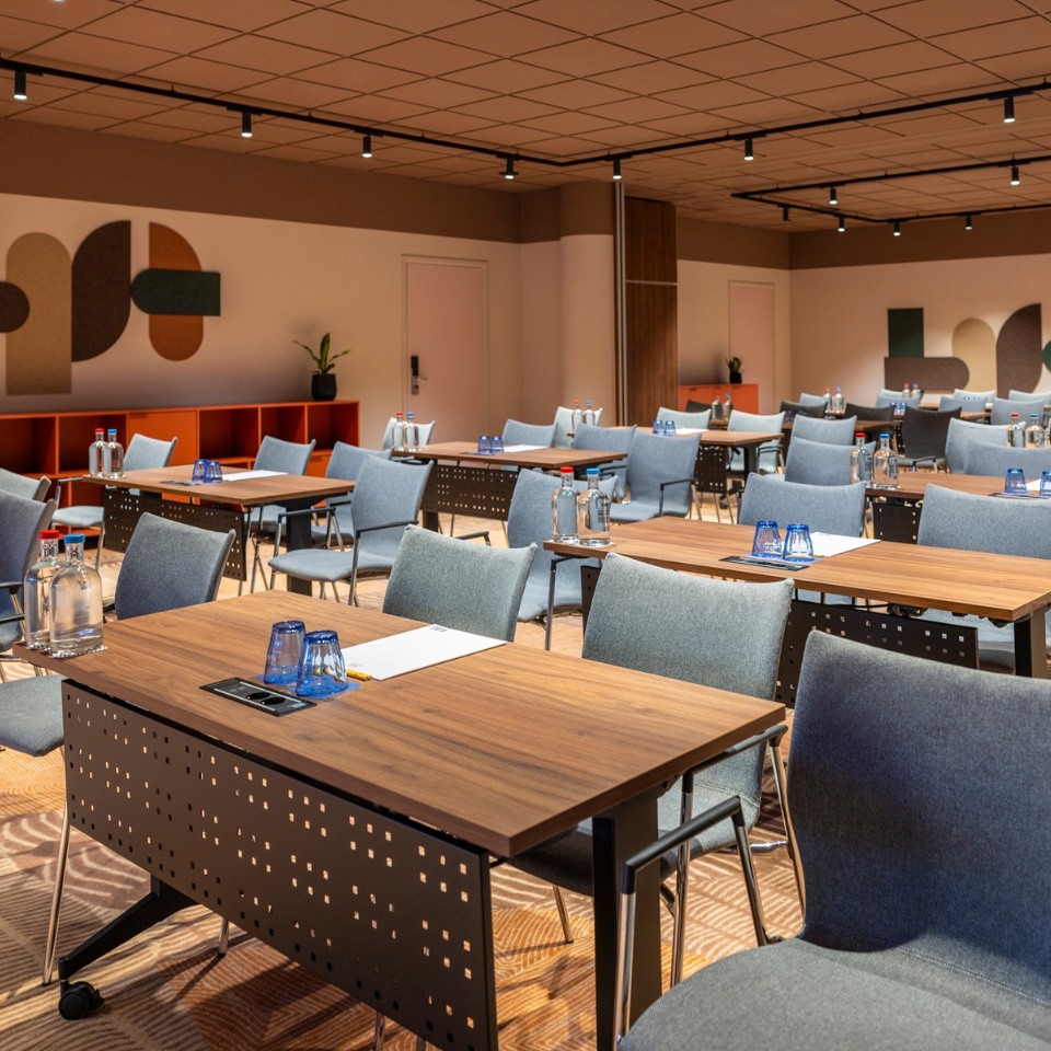 Conference room with modern decor and a warm atmosphere. Wood tables and gray chairs are neatly arranged, with water bottles and glasses set on each table.