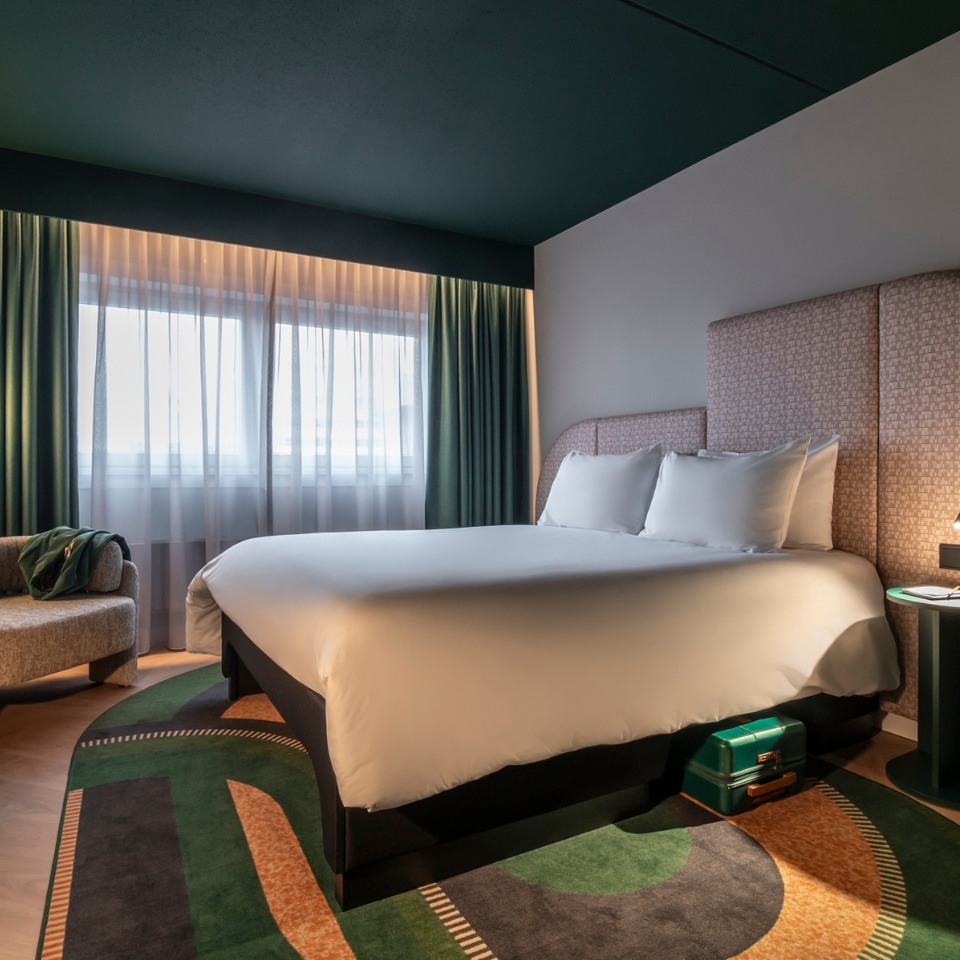 Stylish hotel room with a modern design. Features a large bed with white linens, patterned headboard, soft lighting, green curtains, and a suitcase below.