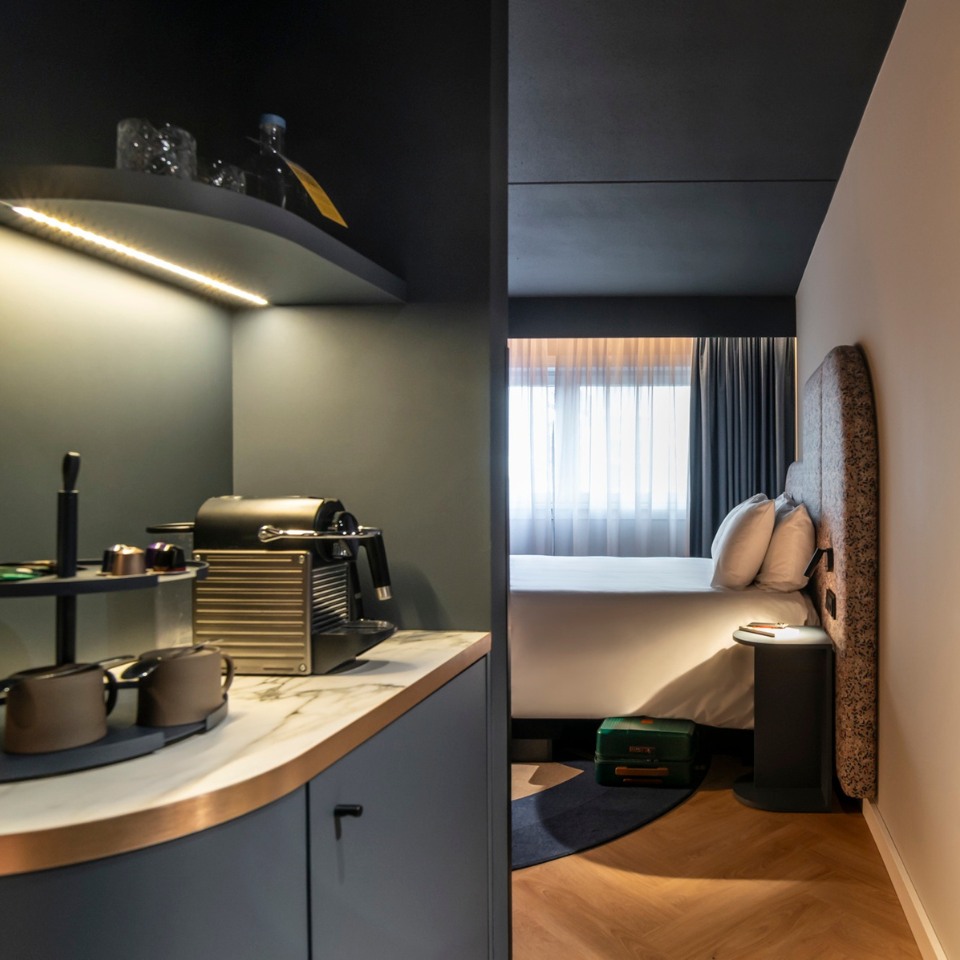 A modern hotel room with a sleek coffee station featuring a coffee maker and cups on the left. The cozy bedroom has soft lighting, plush pillows, and a neatly made bed near a large window, conveying a warm, inviting atmosphere.