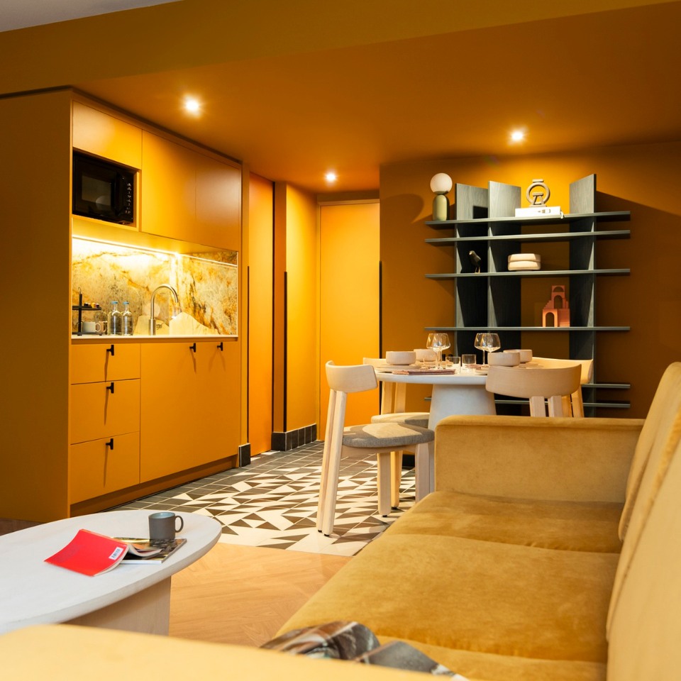Cozy modern kitchen with warm, mustard tones. Features a geometric-patterned floor, yellow cabinets, small dining table, open shelves, and soft lighting.