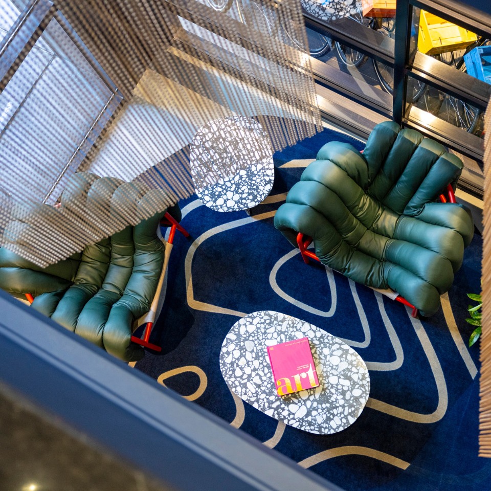 Overhead view of a modern lounge with two green, cushioned chairs on a bold, blue abstract rug. A white patterned table holds a magazine titled "Art."