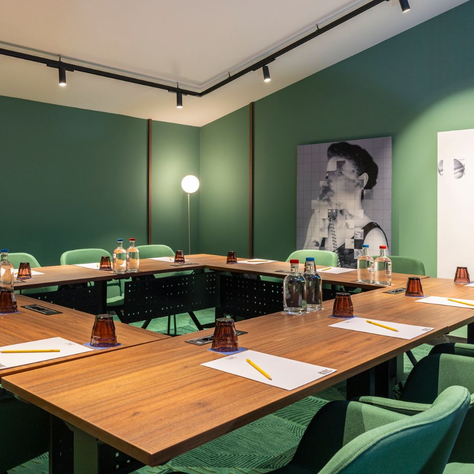 Modern meeting room with green walls and chairs, featuring a U-shaped wooden table set with notepads, pencils, and water bottles. Ambient lighting and abstract art create a stylish atmosphere.