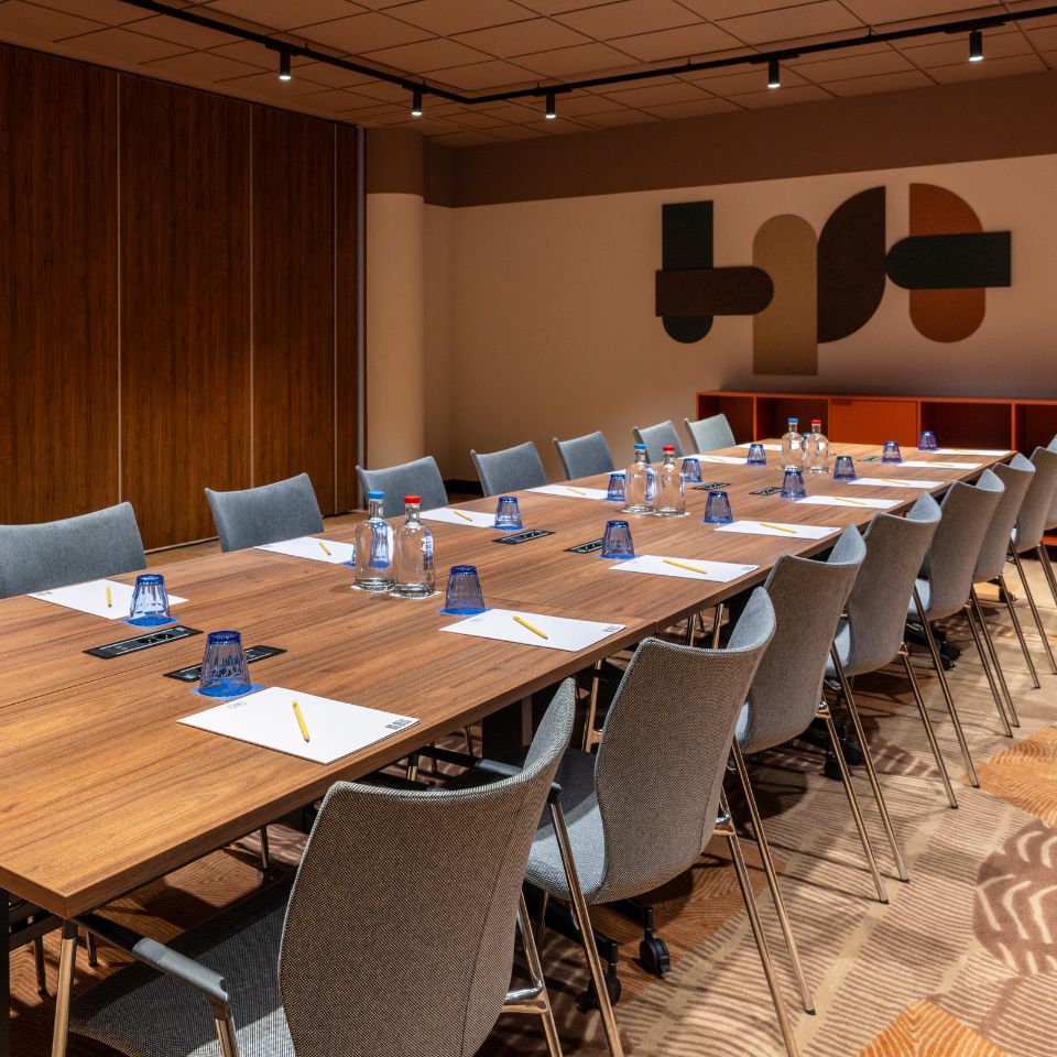 A modern conference room with a long wooden table set with notepads, pencils, water bottles, and blue glasses. Grey chairs surround the table, and abstract art decorates the wall, creating a professional atmosphere.