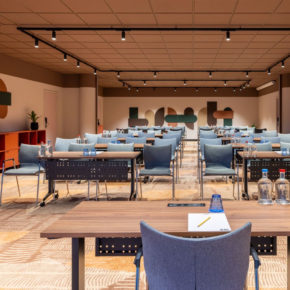 A modern conference room with rows of blue chairs and wooden tables set with notepads and water bottles. Bright lighting and geometric wall art create a professional atmosphere.