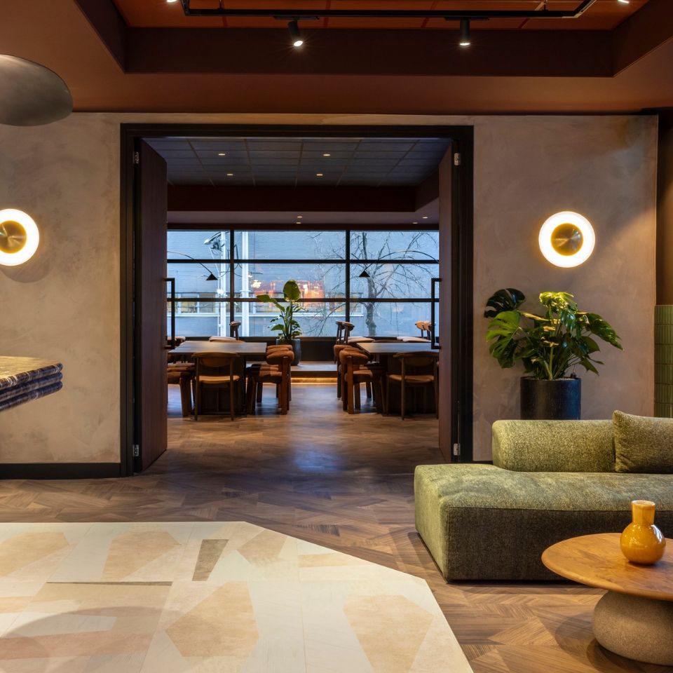 Stylish lounge with green sofa and round table in foreground. Warm lighting, wall plants, and modern decor lead to a cozy dining area with window views.