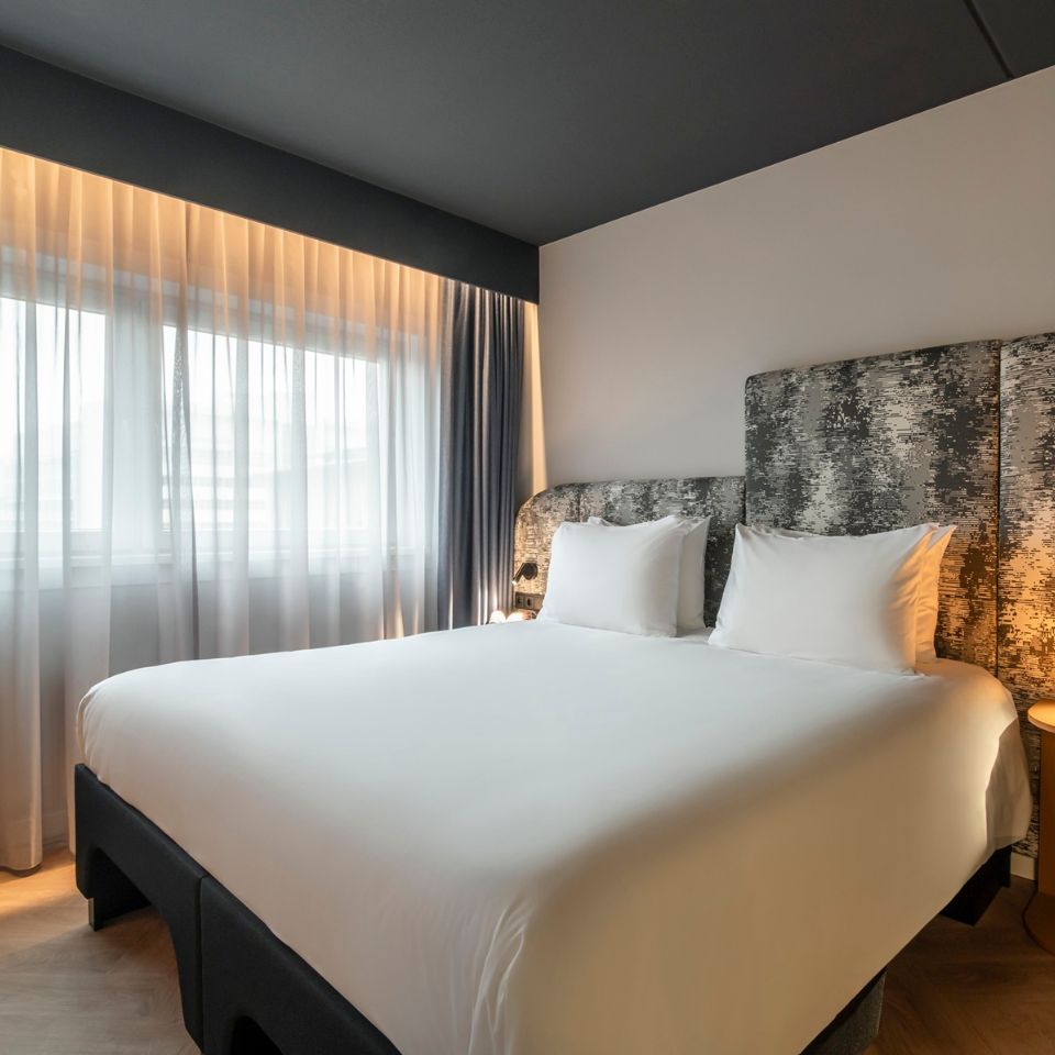 Modern hotel room with a large bed, white linens, and an abstract-patterned headboard. Soft natural light filters through sheer curtains, creating a calm ambiance.