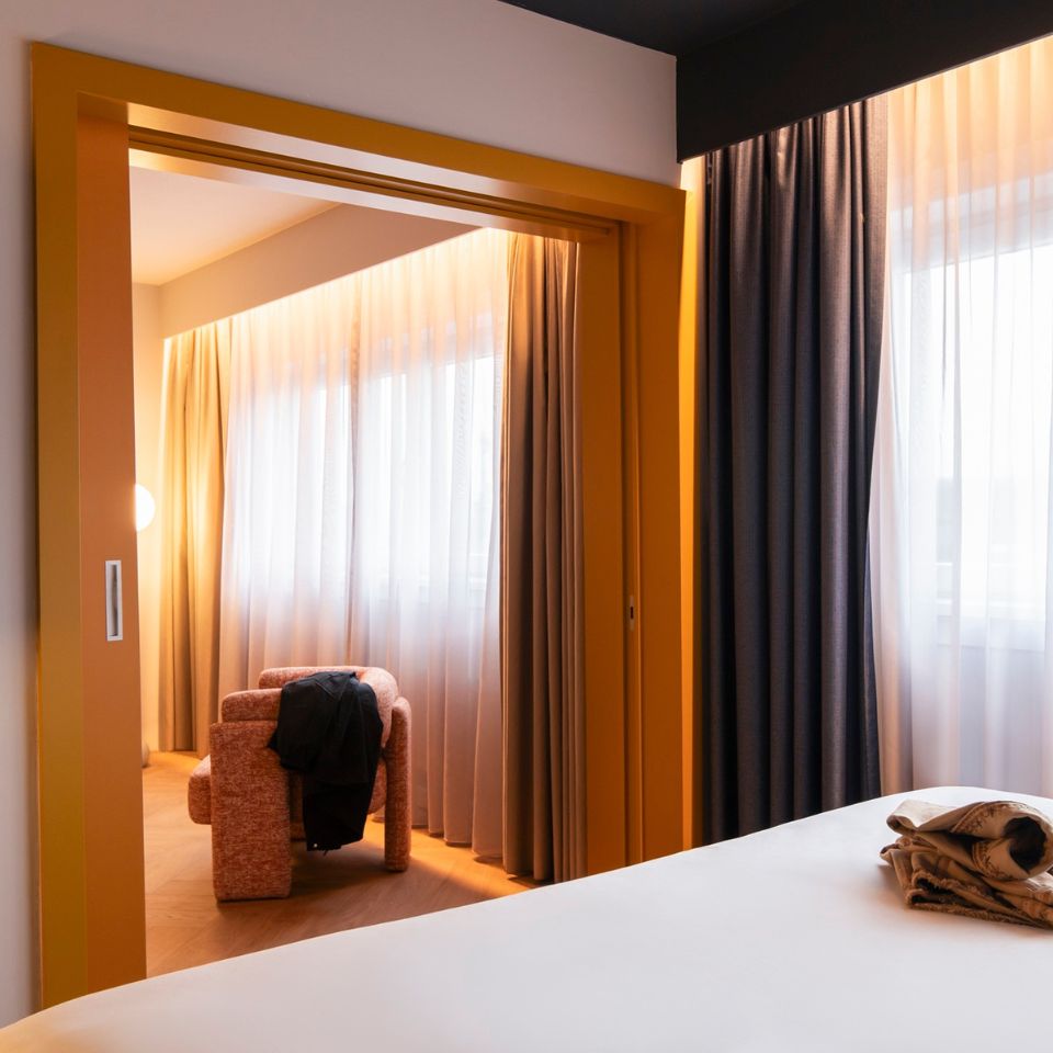 Stylish hotel room with a cozy, peach armchair by large windows with sheer curtains. Soft lighting creates a warm, inviting atmosphere.