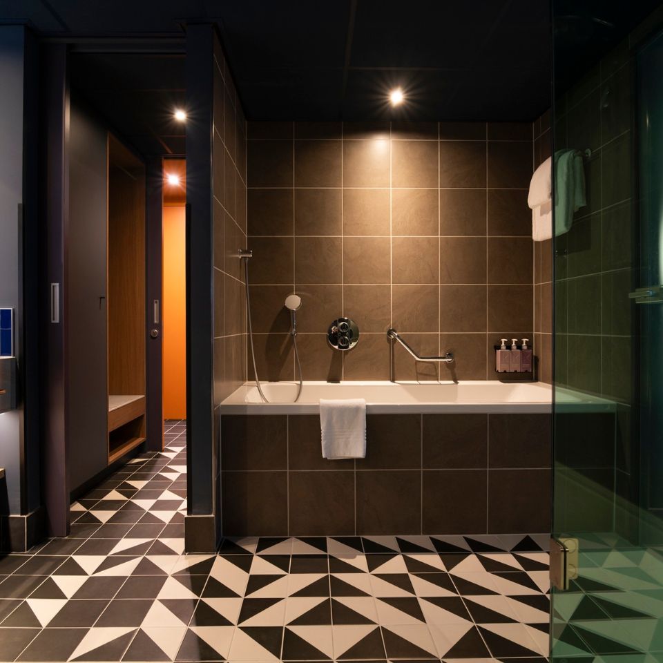 Modern bathroom with geometric tiled floor, brown tiled walls, and a bathtub featuring a handheld shower and faucet. Dim lighting creates a cozy ambiance.