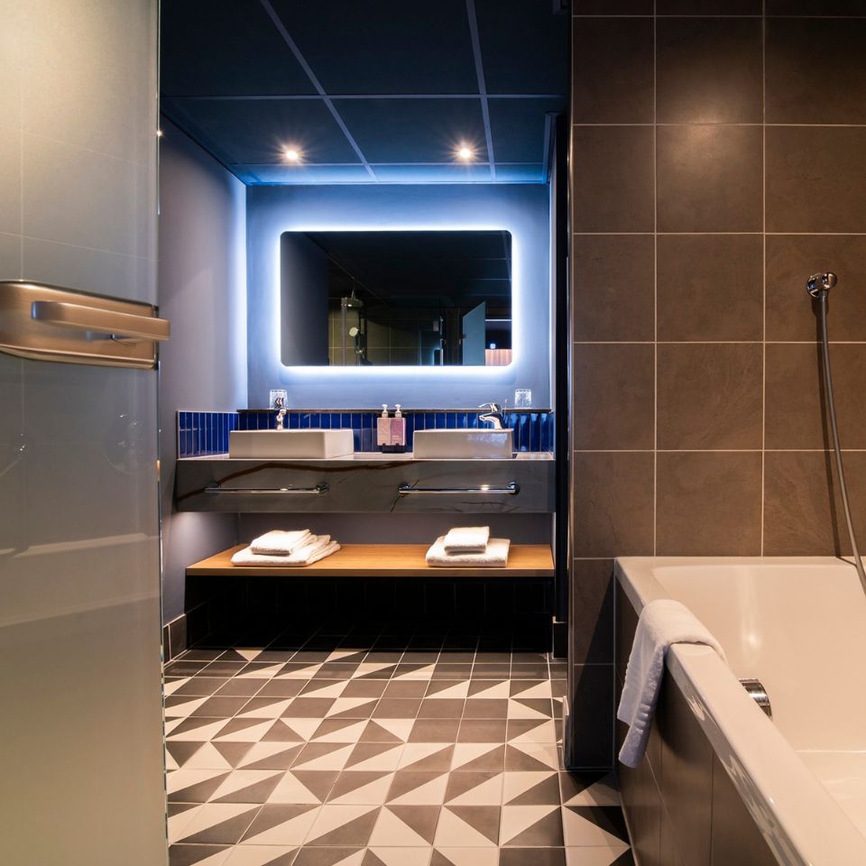 Modern bathroom with geometric floor tiles, a backlit mirror above twin sinks, and a bathtub with a towel. Warm lighting creates a sleek ambiance.