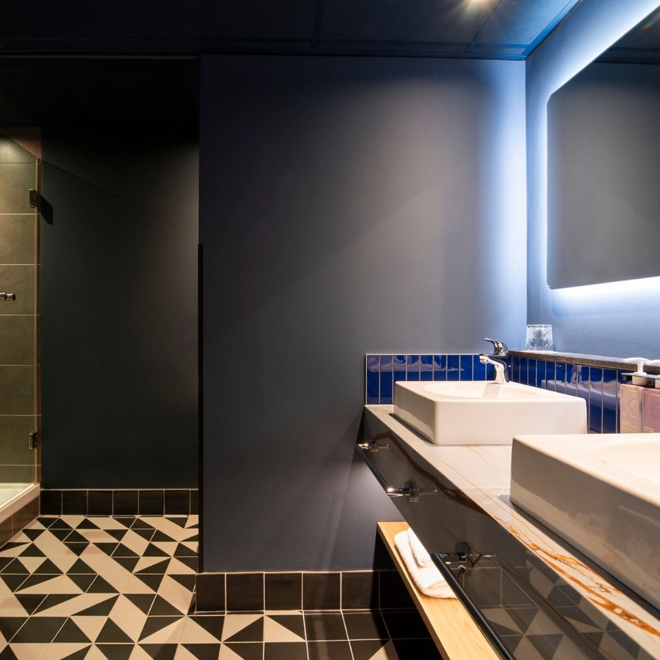 Modern bathroom with geometric tiles, dual sinks with blue backsplash, and a glass shower. Soft lighting creates a sleek, tranquil atmosphere.