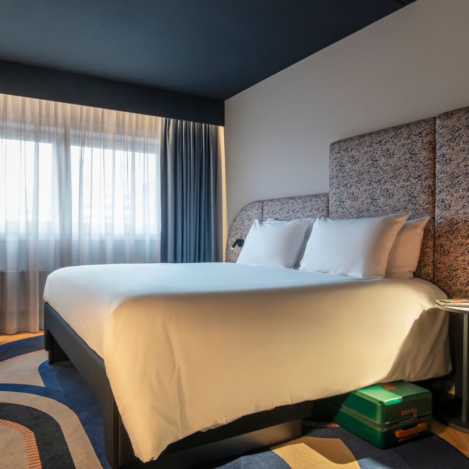 Modern hotel room with a large bed, white linens, and a patterned headboard. Soft, natural light filters through sheer curtains over a window.