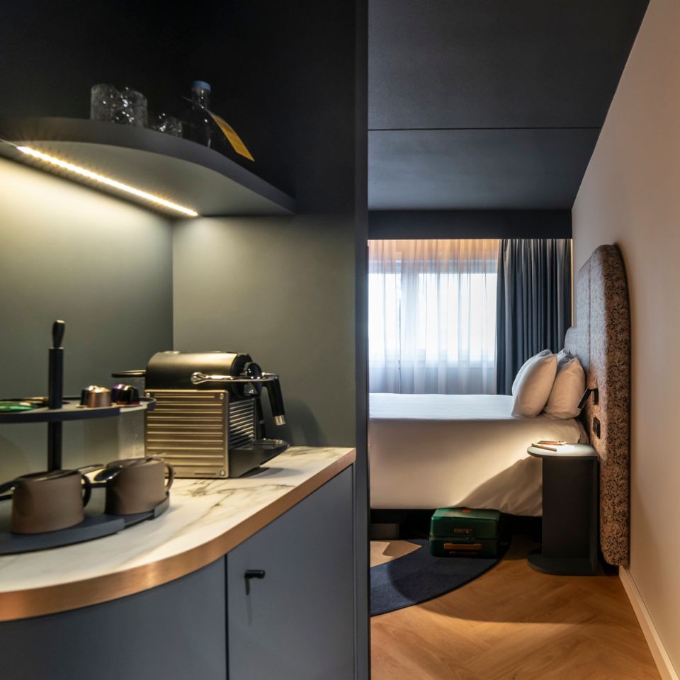Modern hotel room with a cozy atmosphere. Features a marble-topped coffee station with mugs and a machine, leading to a bed by a window with sheer curtains.