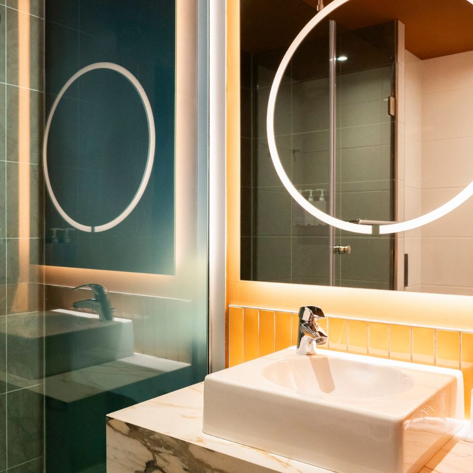 Modern bathroom featuring a square white sink with a sleek silver faucet on a marble counter. A circular, backlit mirror adds a warm, inviting glow.