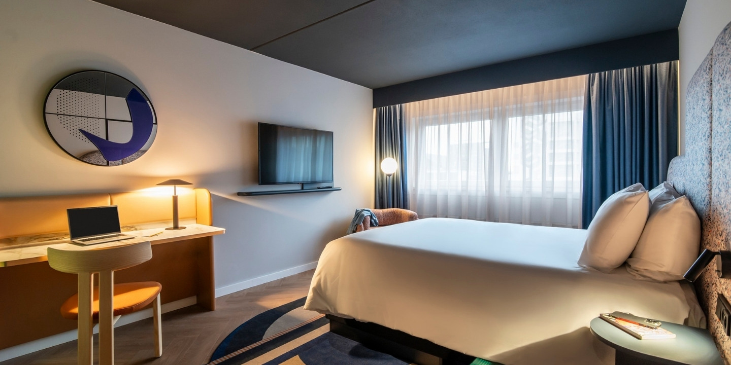 Modern hotel room with a cozy bed, soft lighting, and abstract wall art. Desk with a laptop, TV on the wall, and a window with curtains. Calm ambiance.